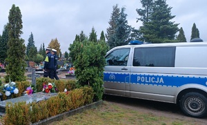 Policjanci ruchu drogowego kontrolują rejon cmentarza. Po lewej stronie zdjęcia widać groby, natomiast po prawej stronie zdjęcia stoi zaparkowany radiowóz policyjny, przed którym widać policjantów.