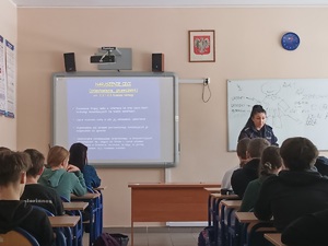 Klasa szkolna w Sypniewie. Policjantka stoi przed uczniami siedzącymi w ławkach szkolnych. Na tablicy interaktywnej wyświetla się prezentacja multimedialna dotycząca zagadnienia nękania.